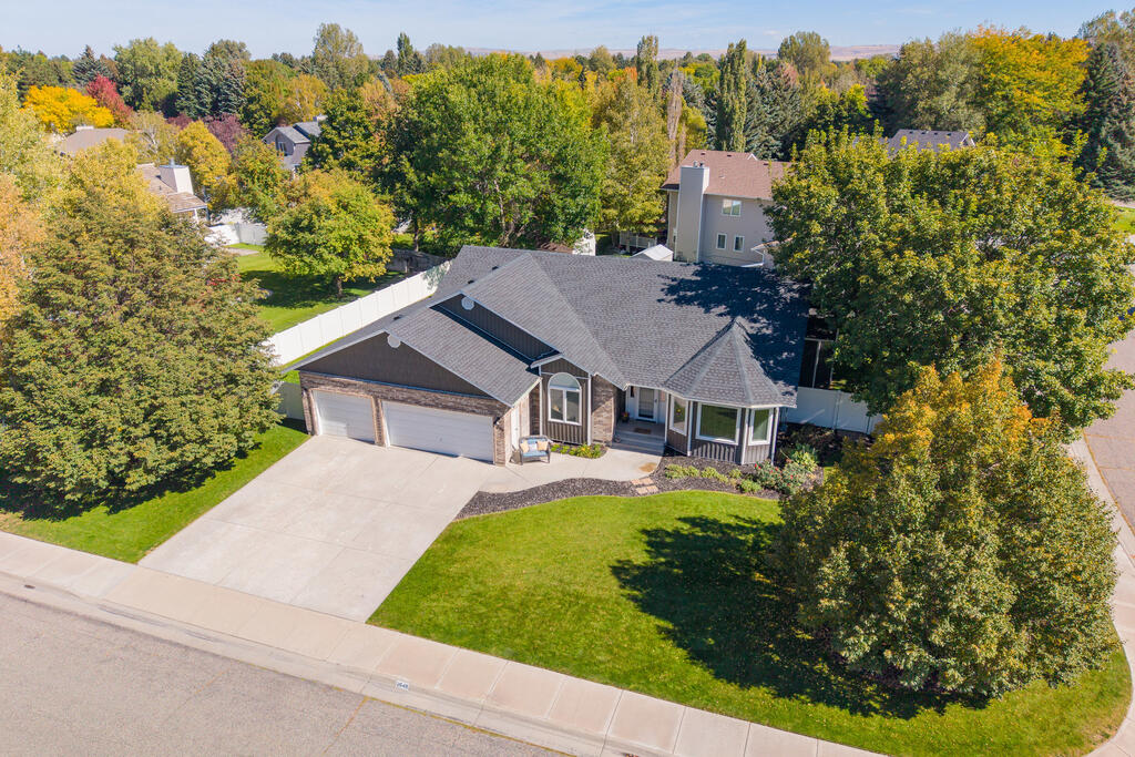 Aerial view of a stone-and-siding home with a wide driveway, front lawn, mature trees, and white fencing.