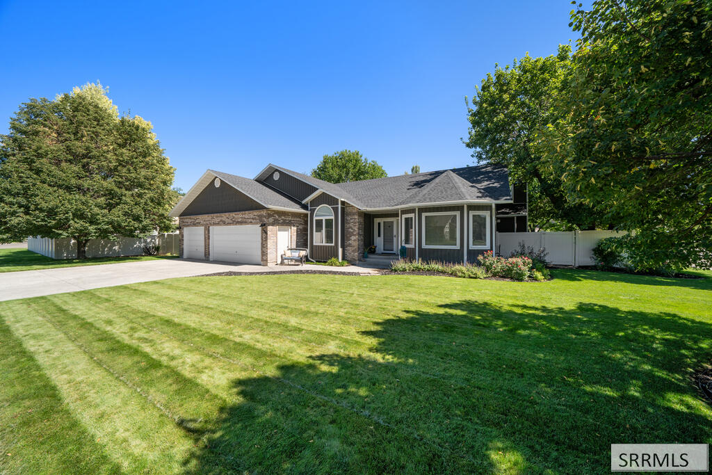 Single-story home with a two-car garage, brick facade, broad green lawn, and mature trees.