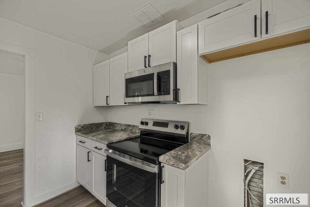 Kitchen with white cabinets, gray stone countertops, and stainless steel appliances; an unfinished wall opening shows exposed wiring.