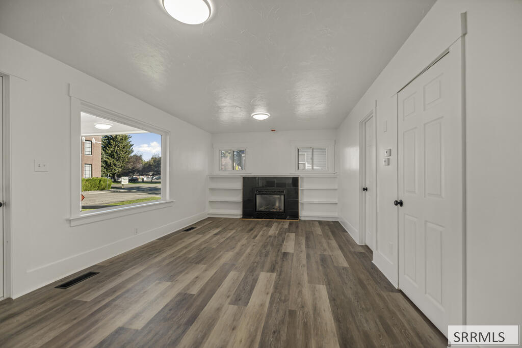 Vacant living room with a fireplace, built-in shelves, gray plank flooring, and a large street-facing window.
