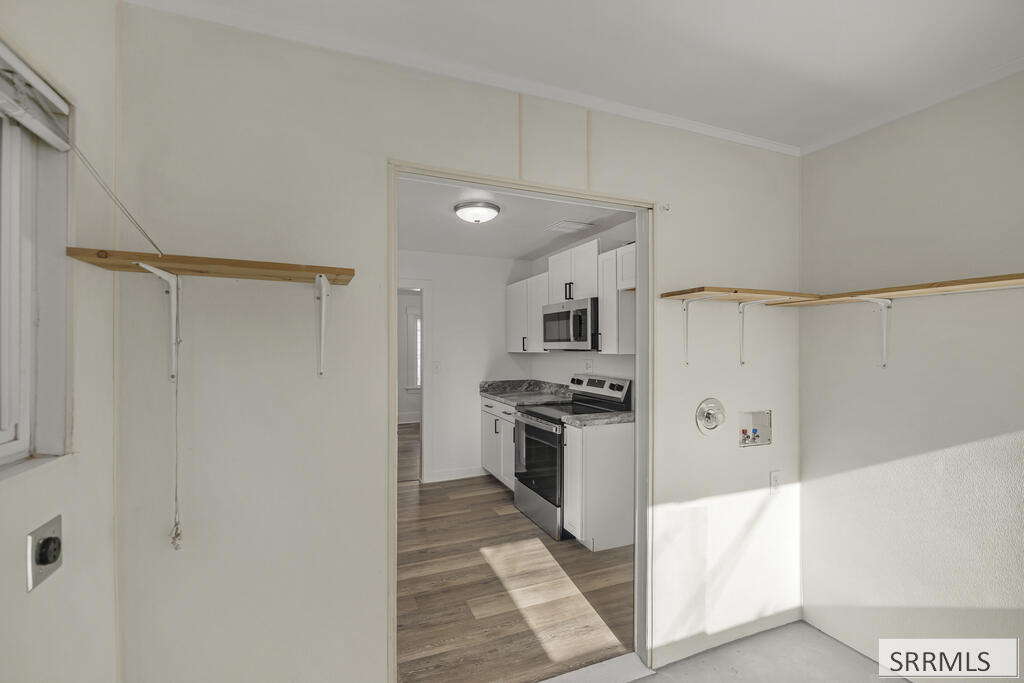 Empty utility room with wall shelves and laundry hookups, opening into a white kitchen.