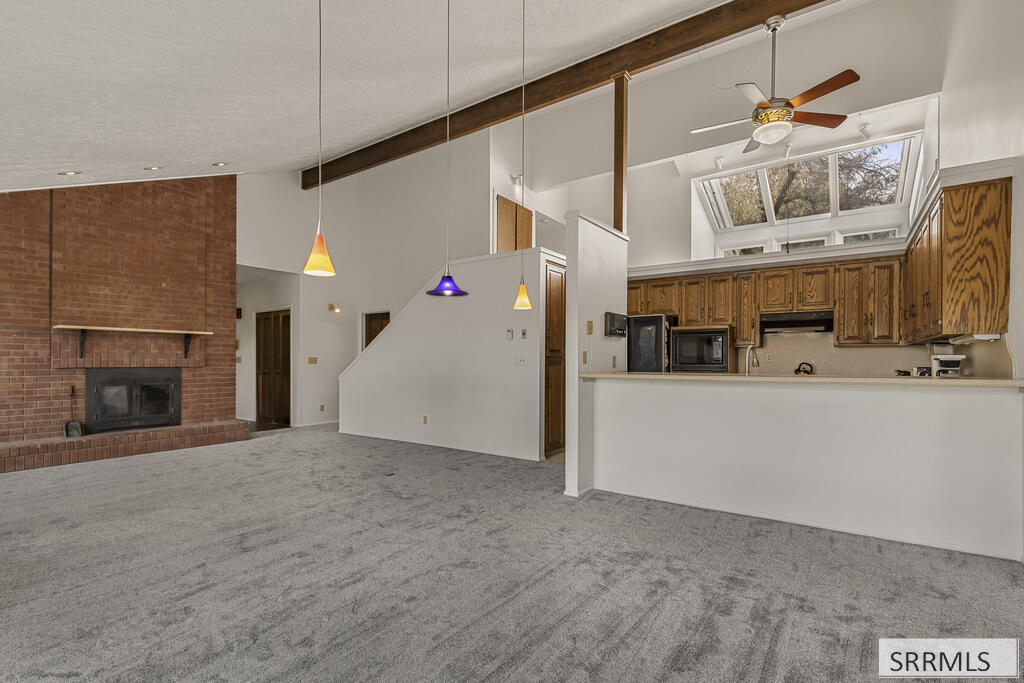 Living room with a brick fireplace, gray carpet, and open kitchen with wood cabinets and high windows.