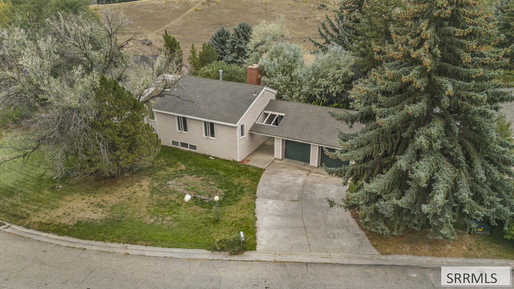 Aerial view of a beige split-level home with a curved driveway, patchy lawn, and mature trees.