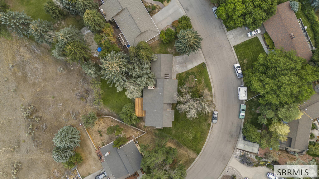 Aerial view of a home among mature trees beside a curved residential street and dry open lot.