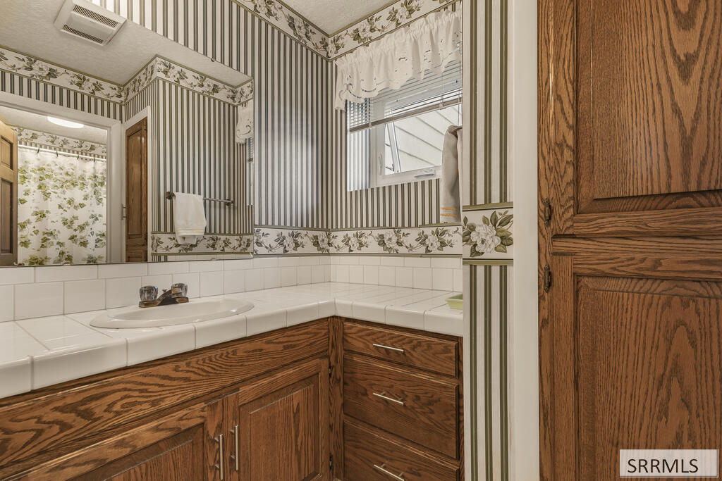 Bathroom vanity with oak cabinets, a tiled countertop, and striped wallpaper with floral trim.