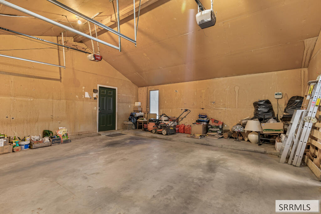 Large garage with a concrete floor, green entry door, lawn equipment, boxes, and ladders.