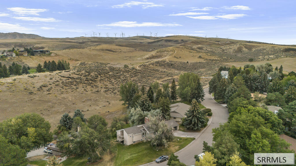 Aerial view of a home beside a curving road, trees, and rolling hills with wind turbines.