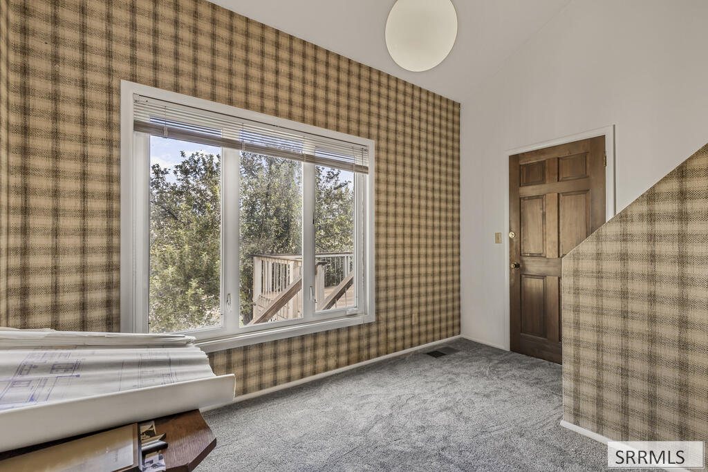 Unfurnished room with plaid wallpaper, gray carpet, and a large window overlooking trees and a deck.