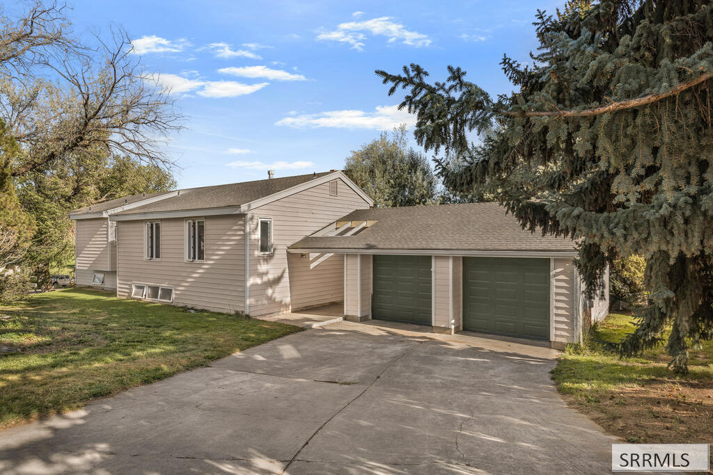 Beige-sided house with an attached two-car garage and wide concrete driveway, framed by mature trees.