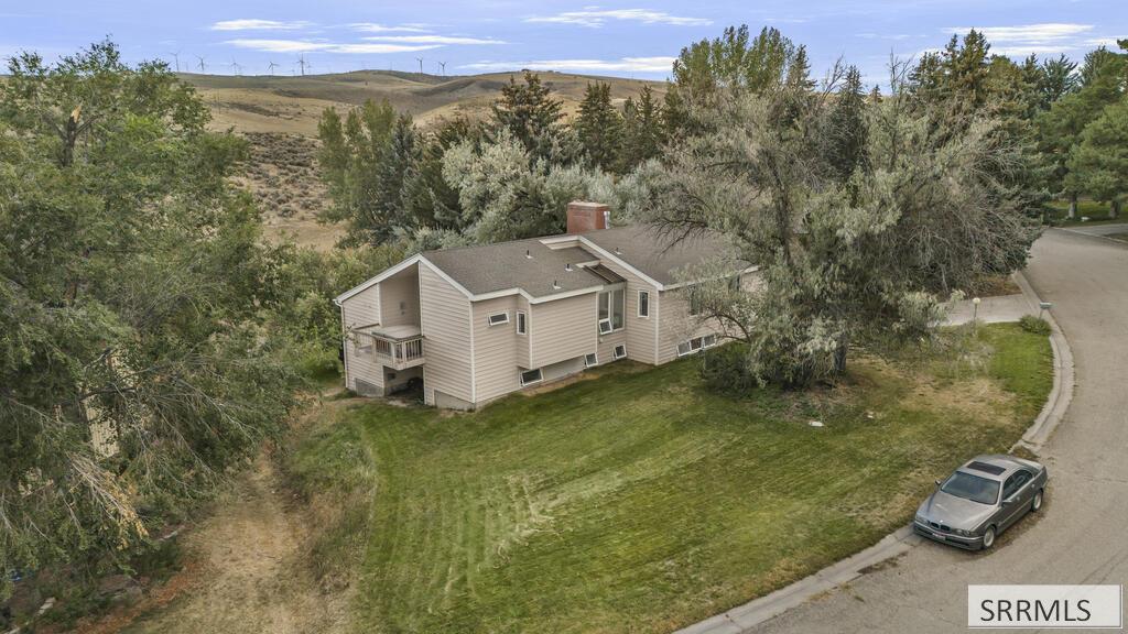 Beige multi-level home with a small deck, broad lawn, mature trees, and distant wind turbines.
