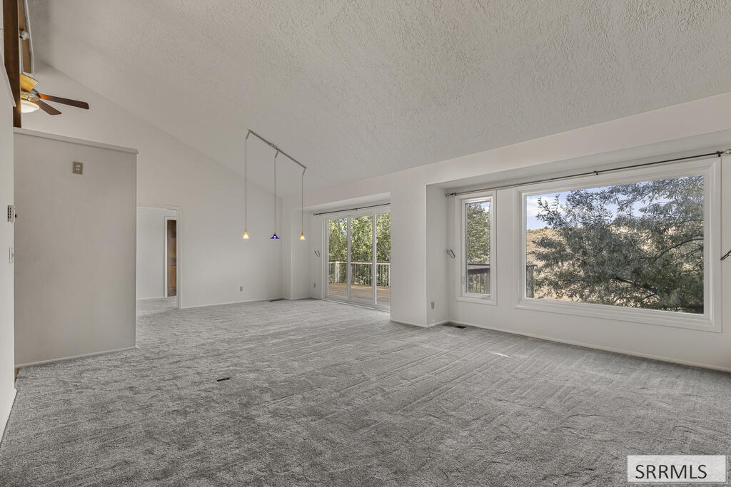 Empty living room with gray carpet, pendant lights, and large windows overlooking trees.