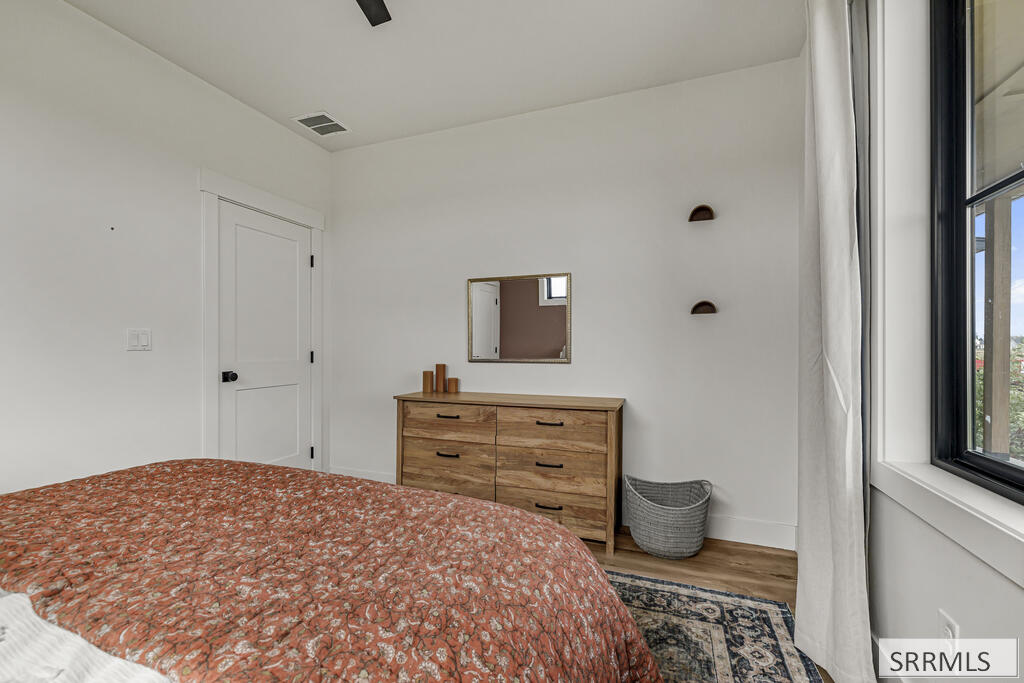 Bedroom with a rust floral quilt, wooden dresser, patterned rug, and large curtained window.