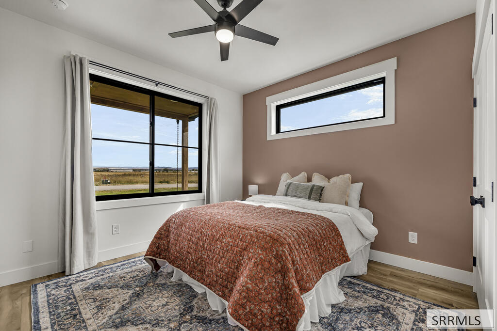 Staged bedroom with a rust-colored quilt, brown accent wall, and windows overlooking open fields.