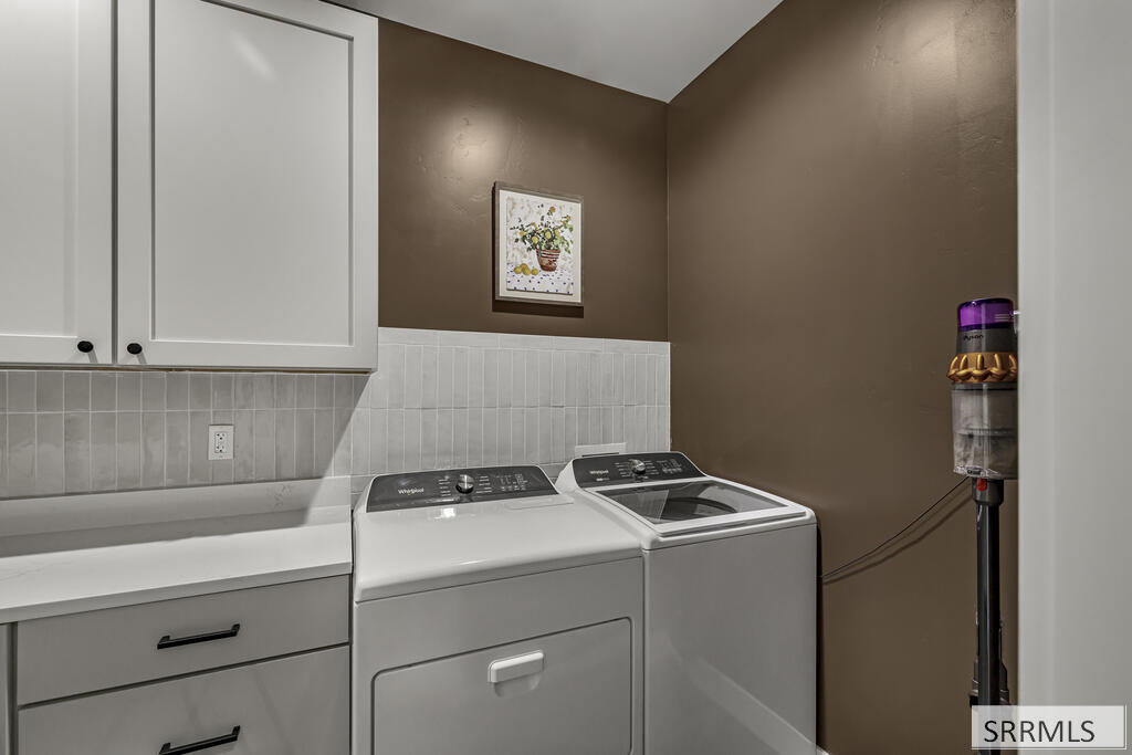 Laundry room with white washer and dryer beneath cabinets and a tiled backsplash.