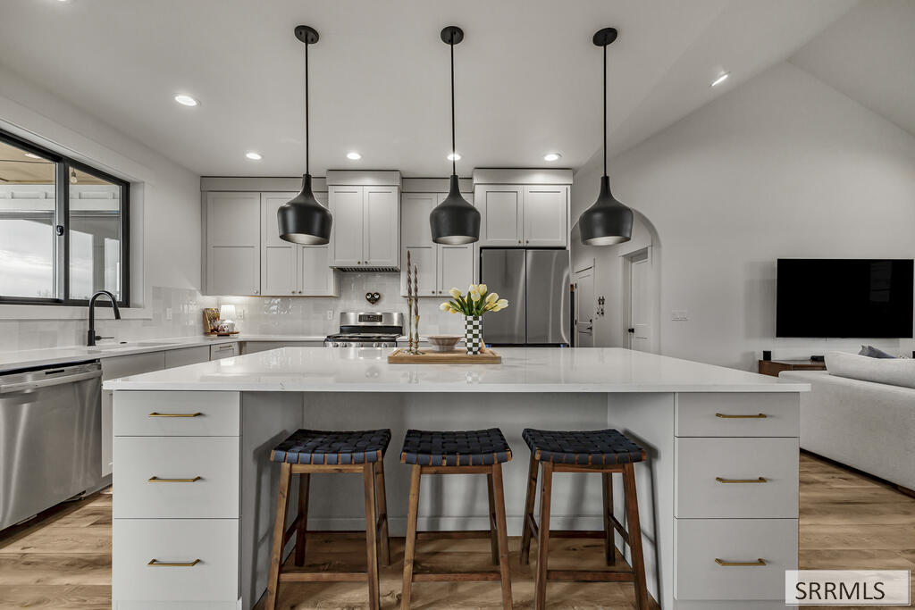 Modern white kitchen with a large island, three stools, black pendant lights, and stainless steel appliances.