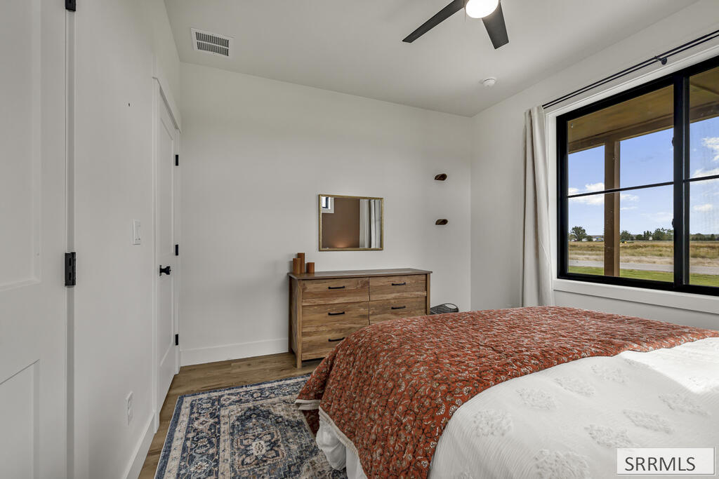 Bedroom with a rust-patterned quilt, wooden dresser, and large window overlooking open fields.