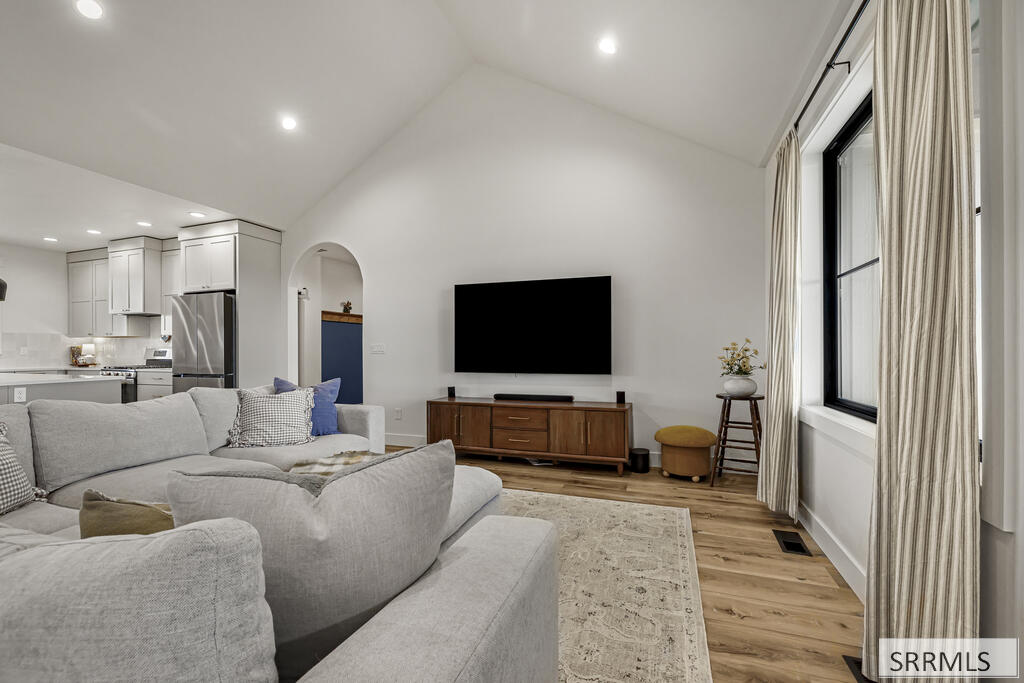 Living room with vaulted ceiling, sectional sofa, and large curtained window beside an open kitchen.