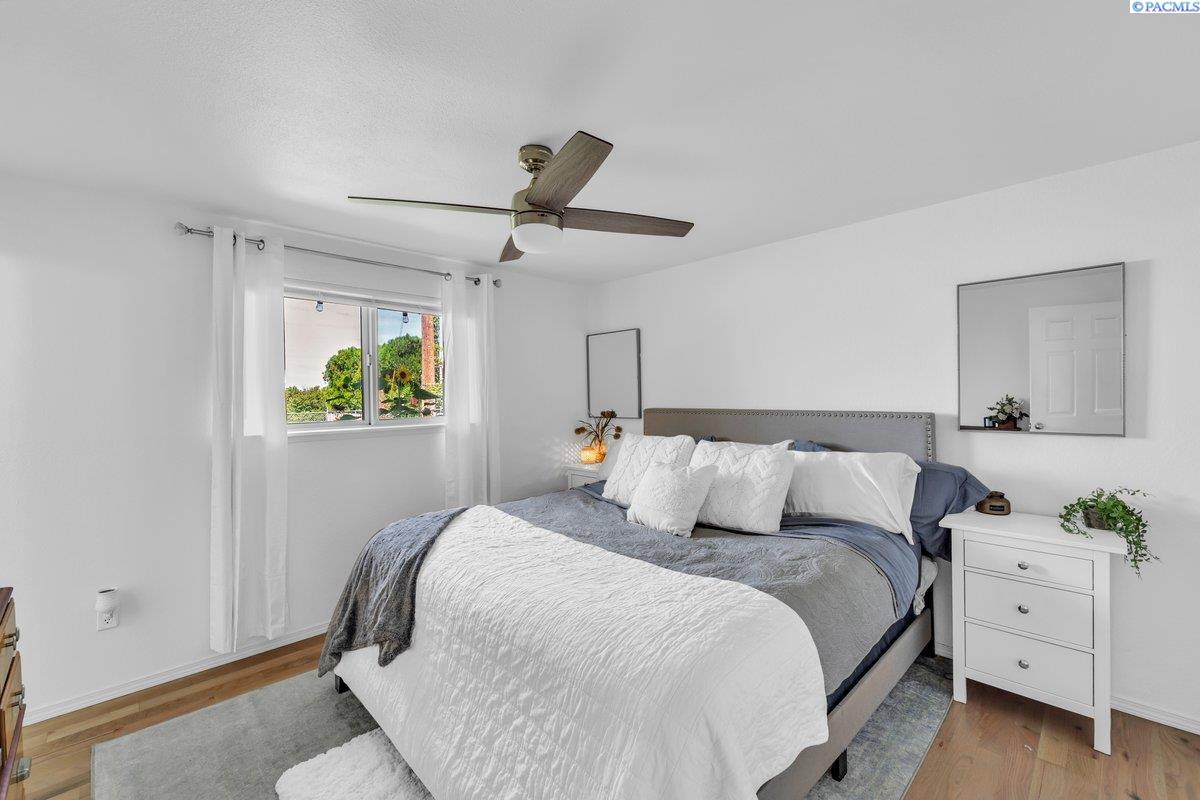 Furnished bedroom with a large gray-and-white bed, ceiling fan, and window with white curtains.