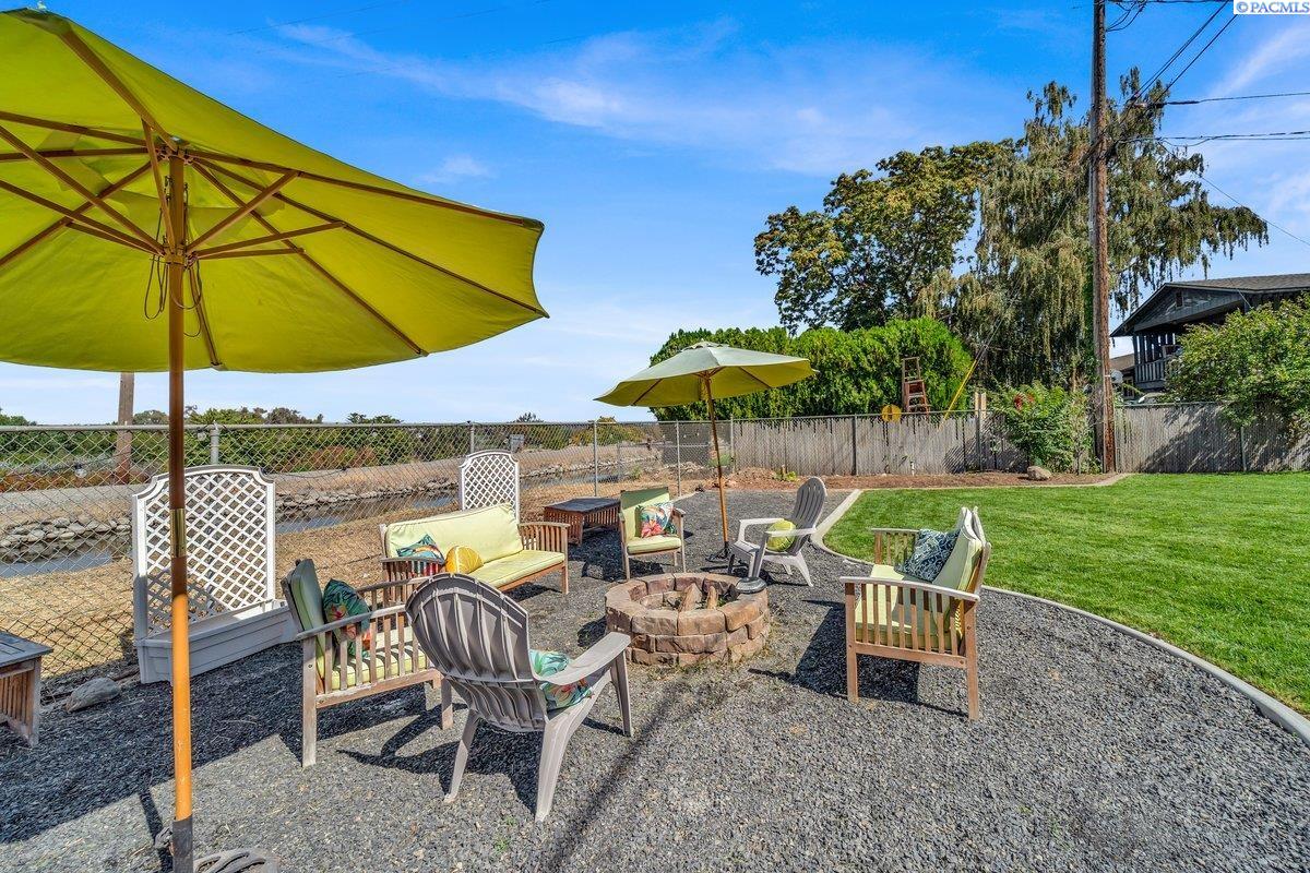 Backyard patio with umbrella-shaded seating, a stone fire pit, and lawn beside a fenced waterway.