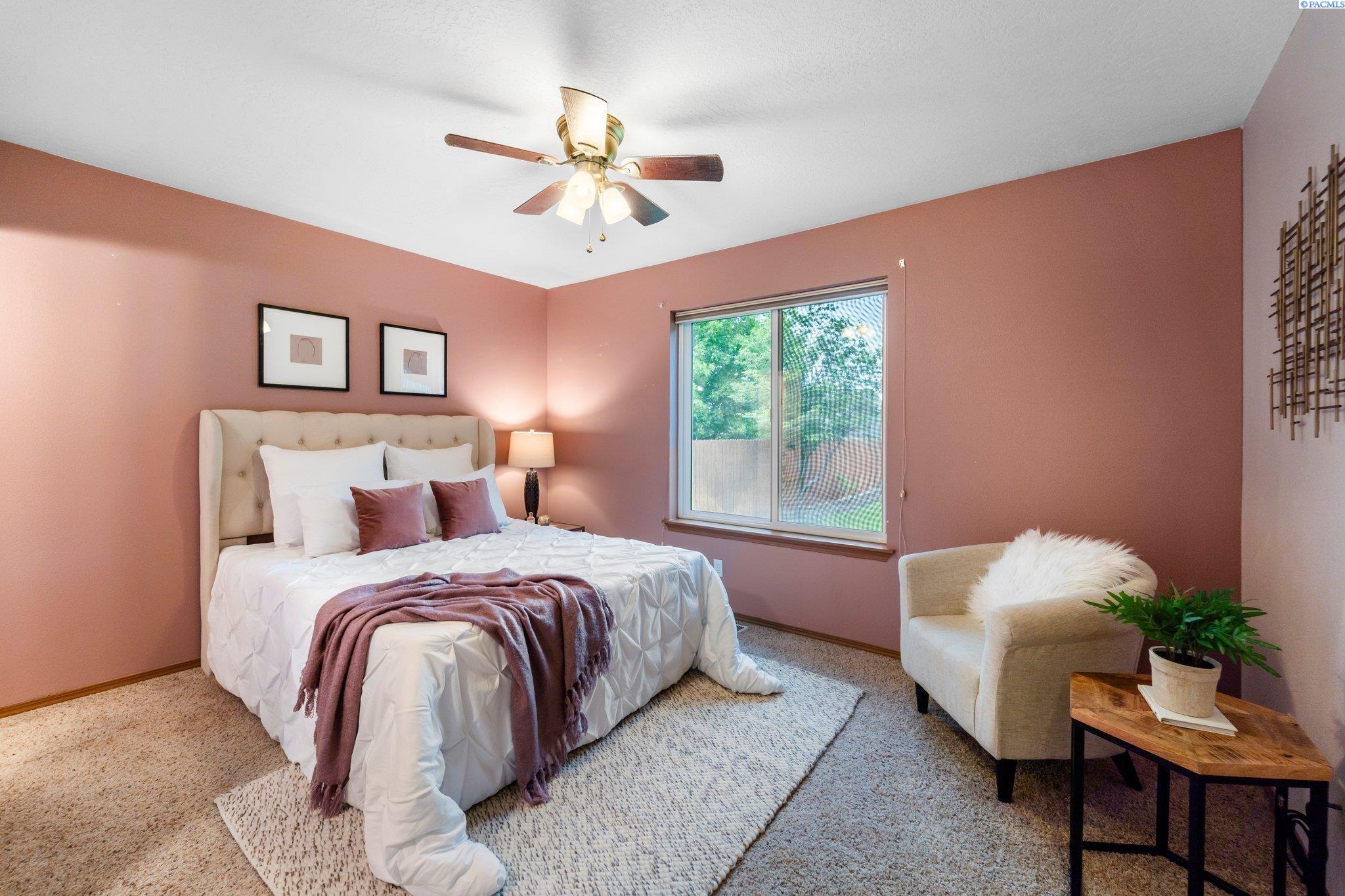 Staged bedroom with a cream upholstered bed, mauve walls, and a cream armchair by the window.