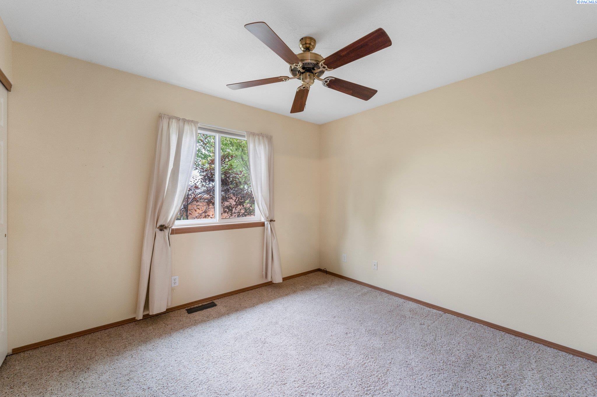 Empty bedroom with beige walls, carpet, a wood ceiling fan, and a curtained window.