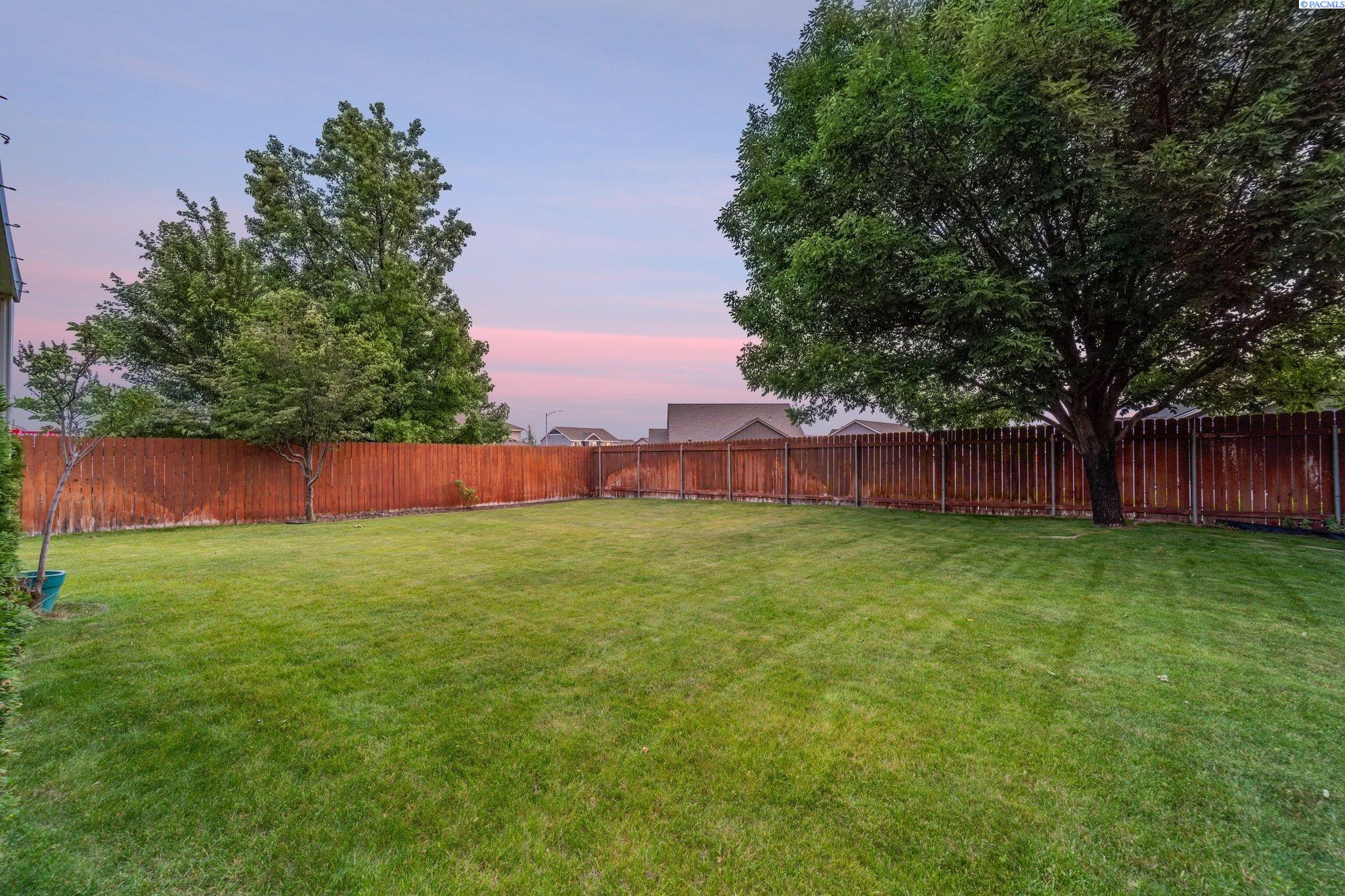 Fenced backyard with a broad lawn, mature shade trees, and a pink dusk sky.