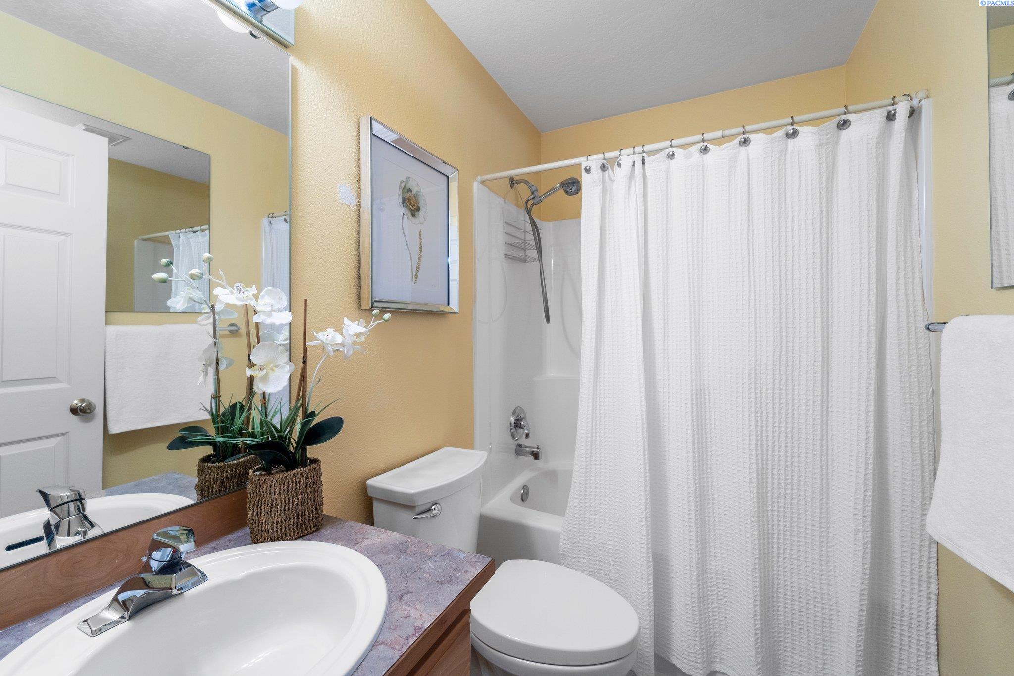 Bathroom with a white sink, mauve countertop, toilet, and curtained tub; white orchids sit beside the mirror.