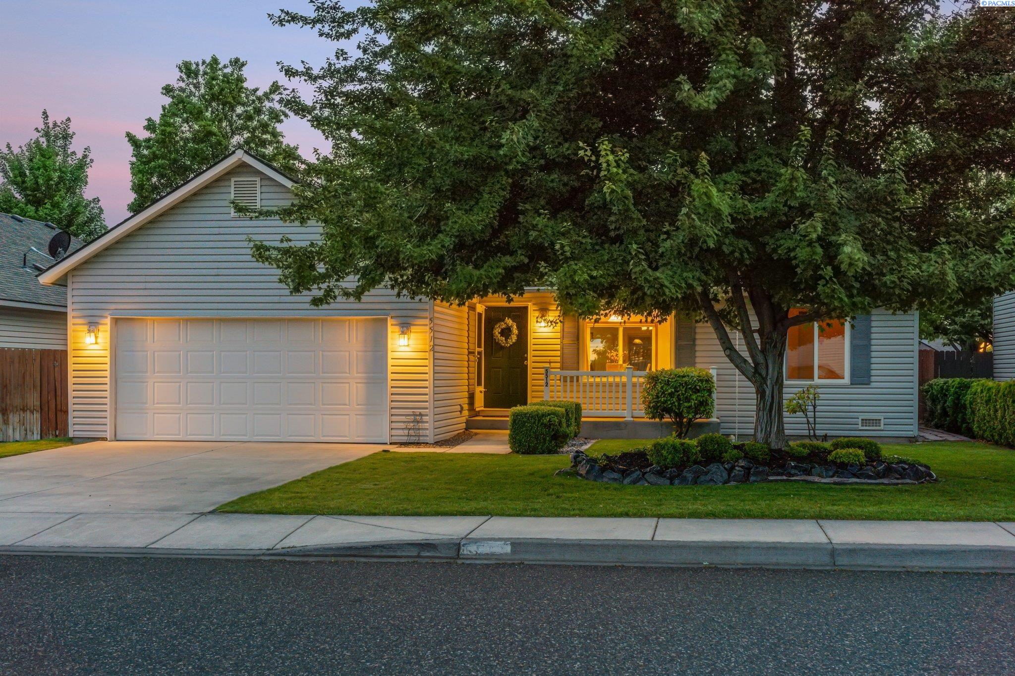 House at dusk with a two-car garage, warmly lit porch, and mature tree in the front yard.