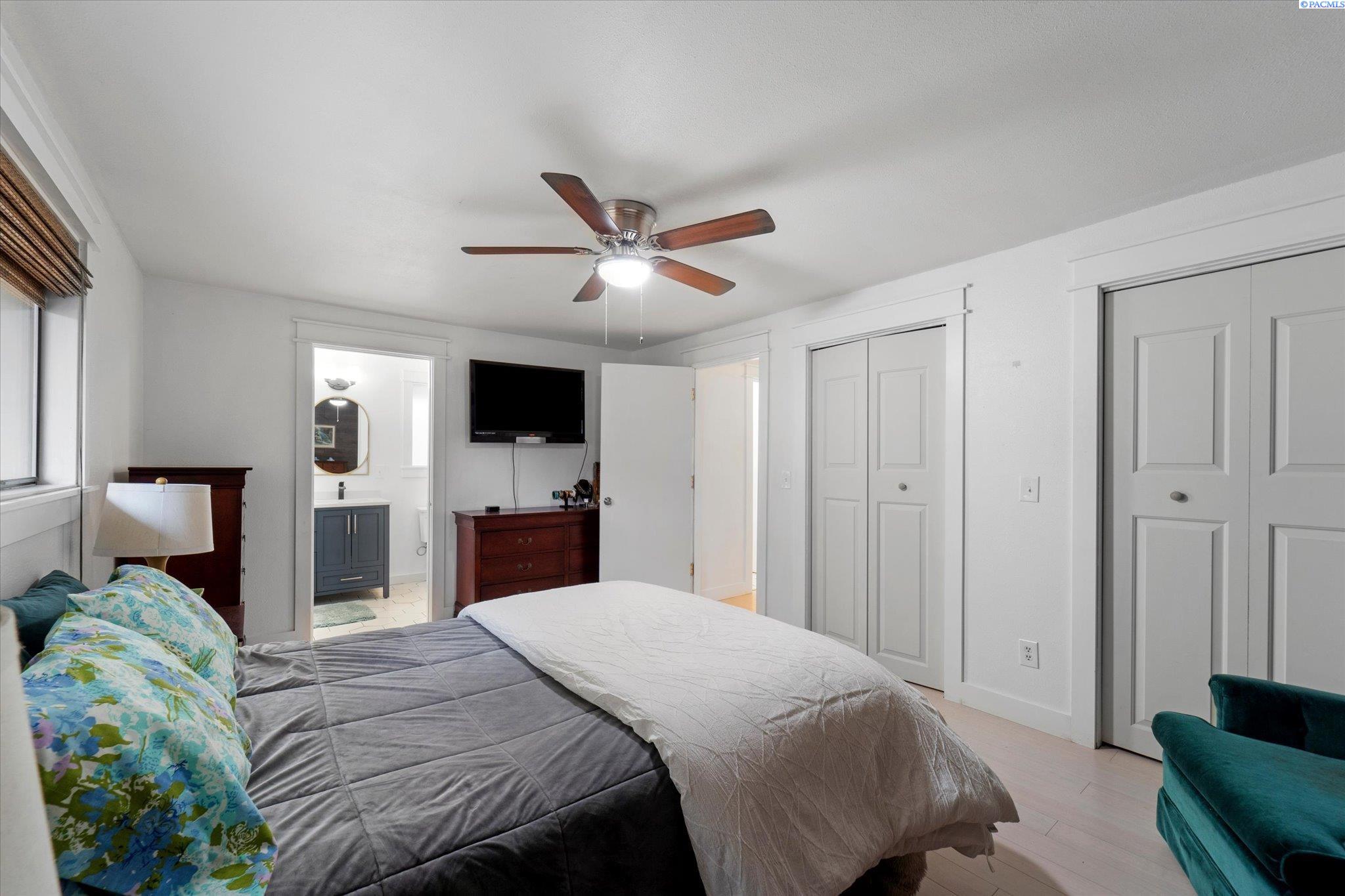 Bedroom with a ceiling fan, wall-mounted television, and open doorway to a bathroom.