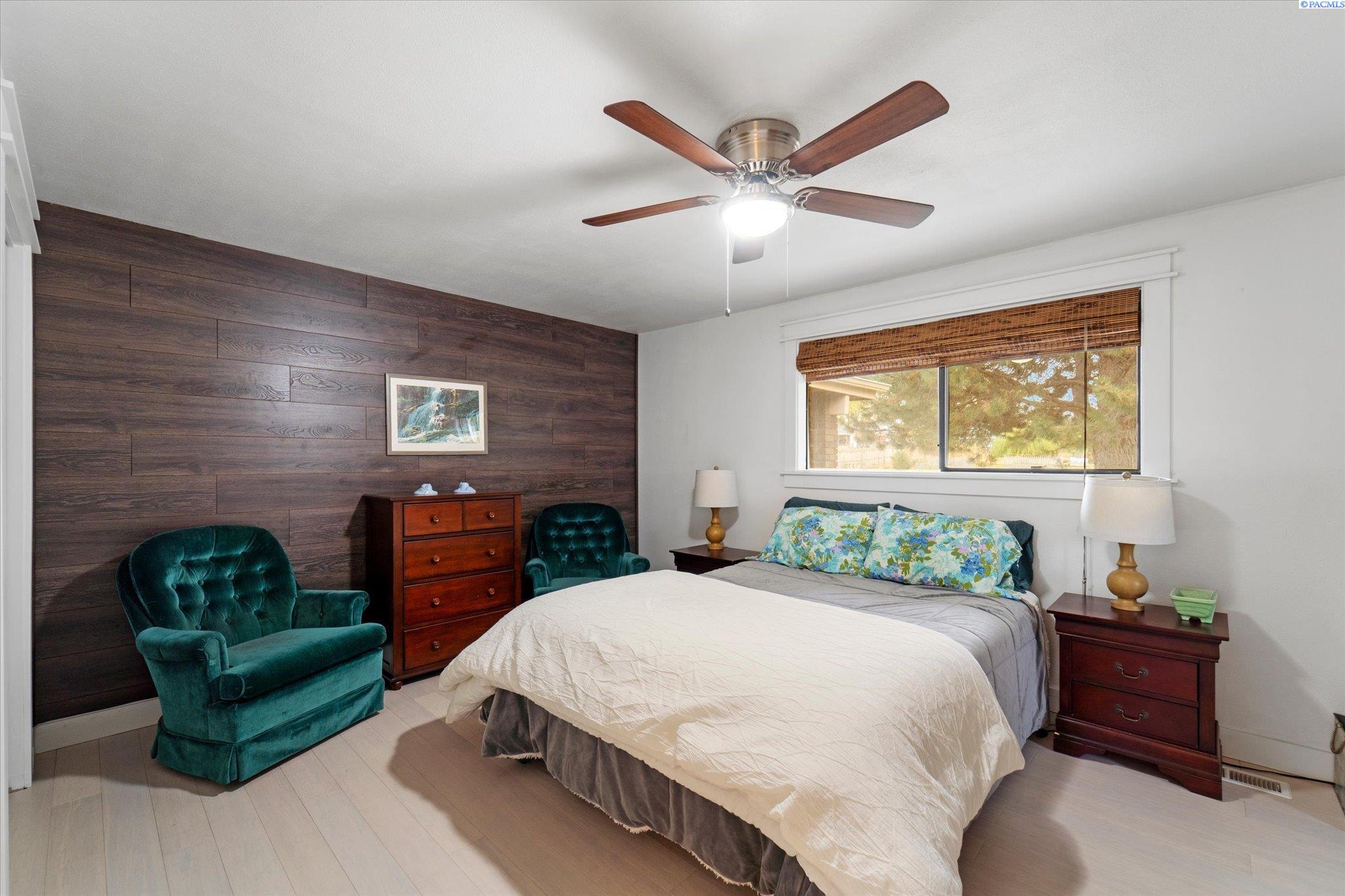 Furnished bedroom with a dark wood accent wall, teal chairs, and a ceiling fan.