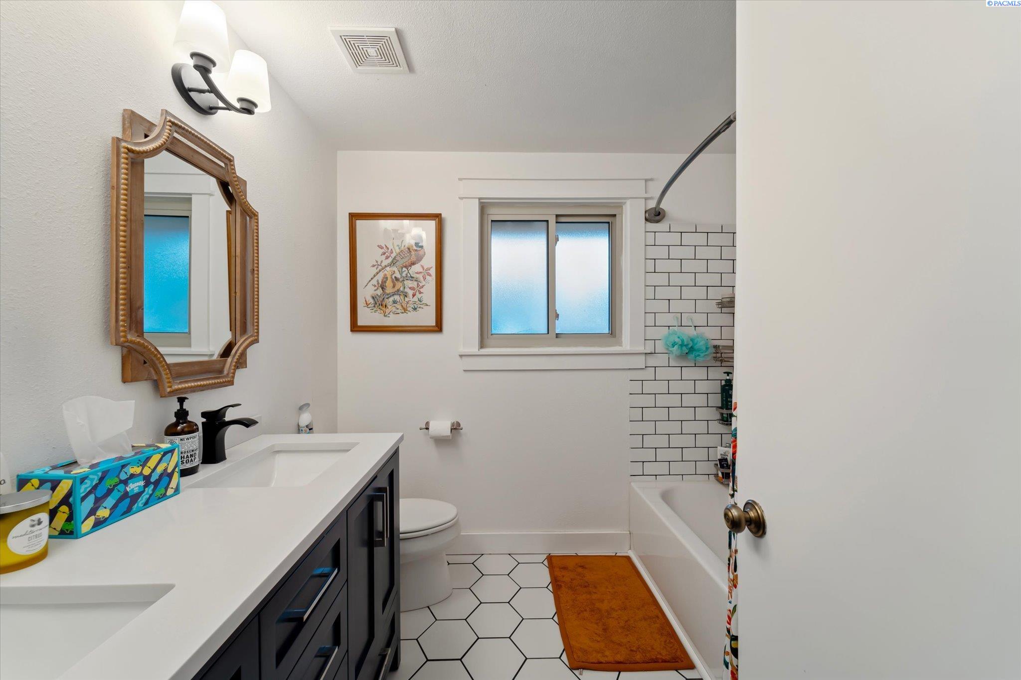 Bathroom with a dark vanity, ornate mirror, frosted window, and white hexagonal floor tile.