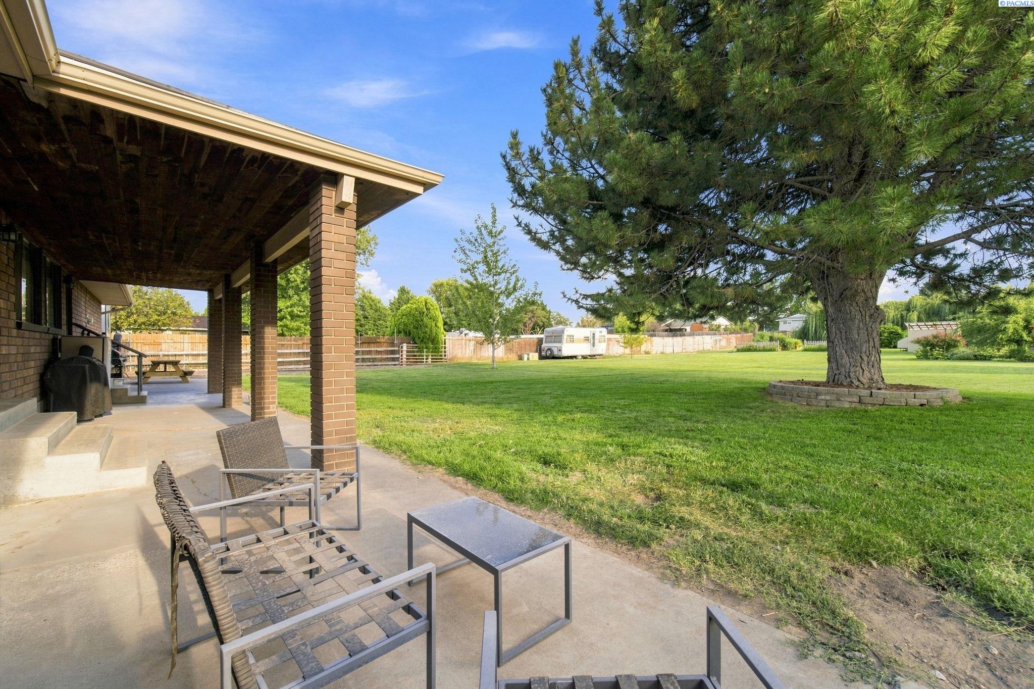 Covered brick patio with outdoor seating beside a spacious lawn and mature pine tree.