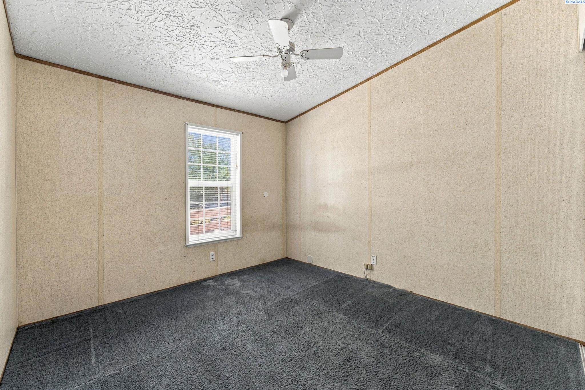 Empty carpeted room with beige walls, a window with blinds, and a ceiling fan.