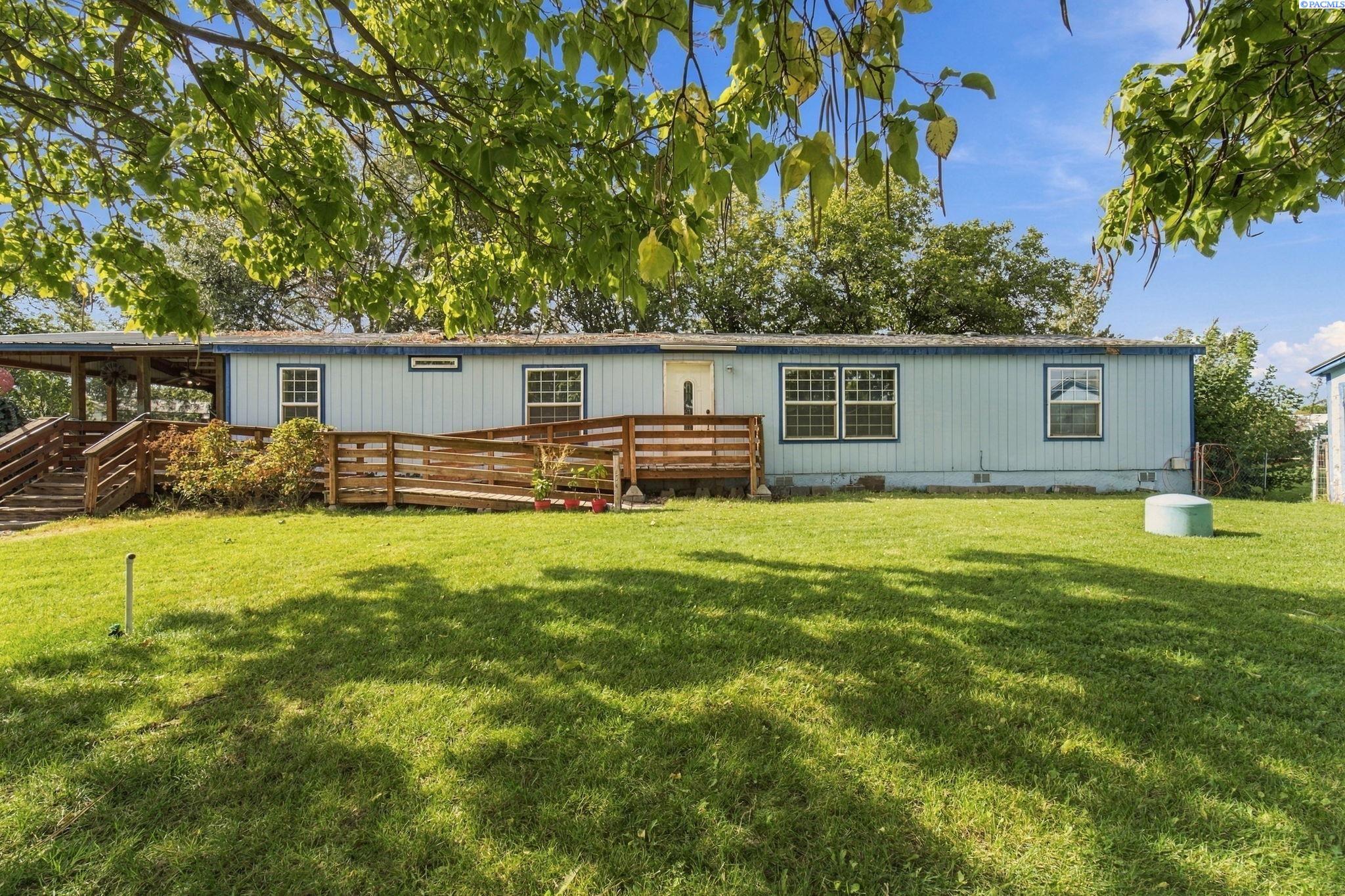 Light-blue single-story home with a wooden ramp and deck beneath mature shade trees.