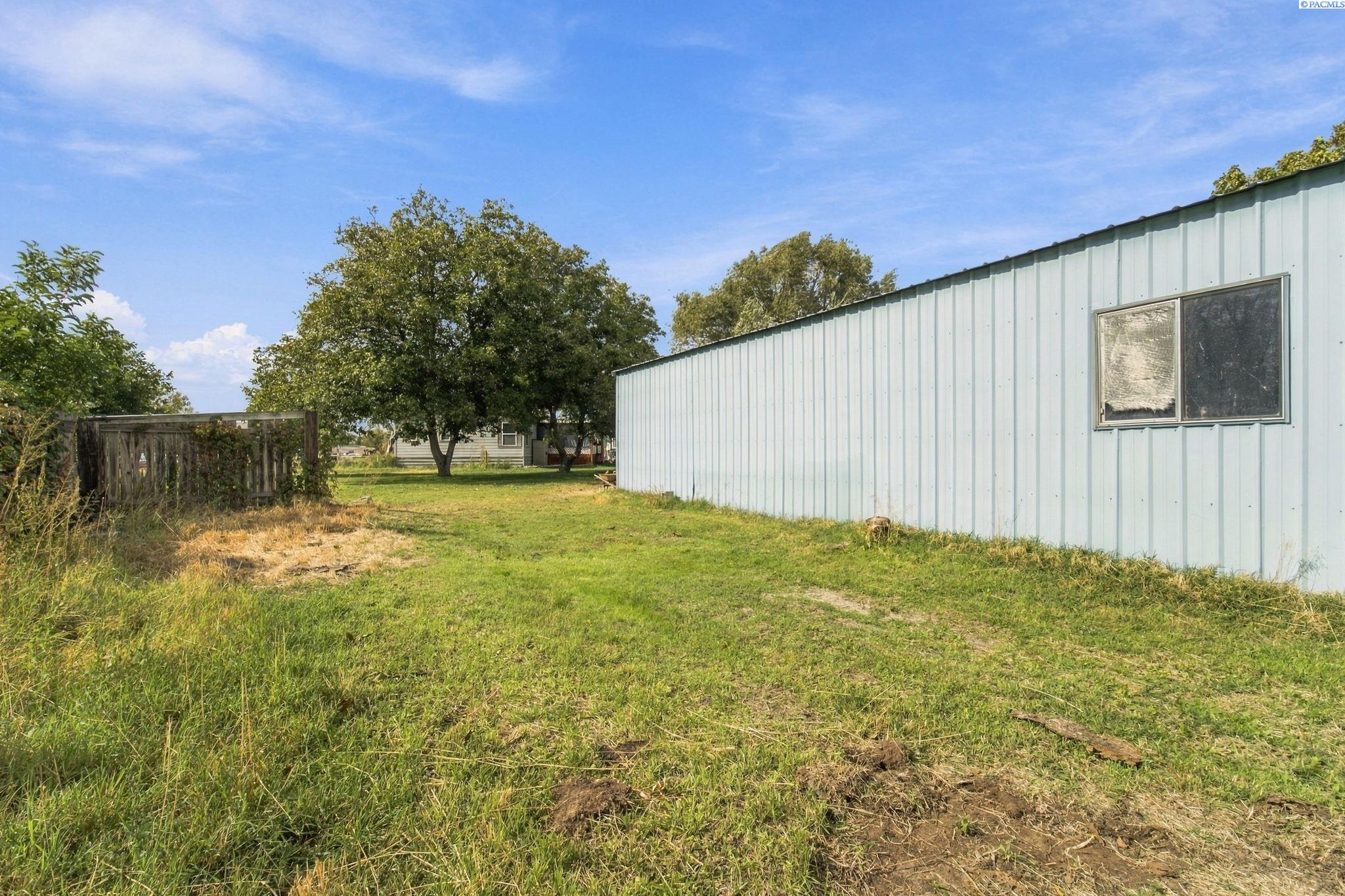 Side yard beside a light metal-sided building, with patchy grass, trees, and a weathered wooden fence.