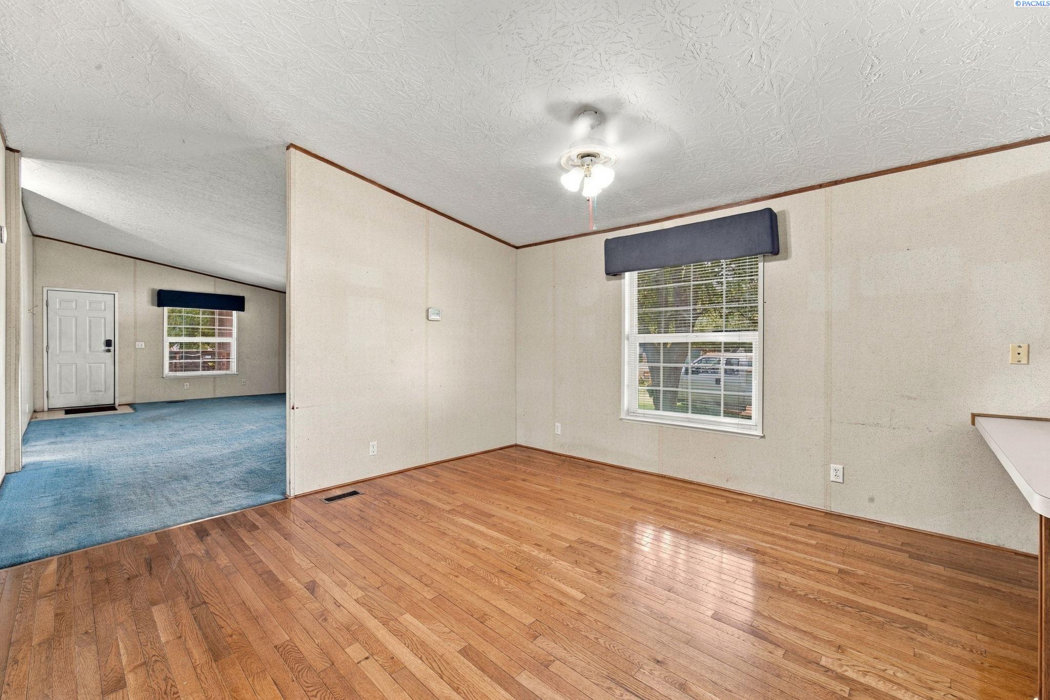 Empty room with hardwood floors and a window, opening onto a blue-carpeted living area.