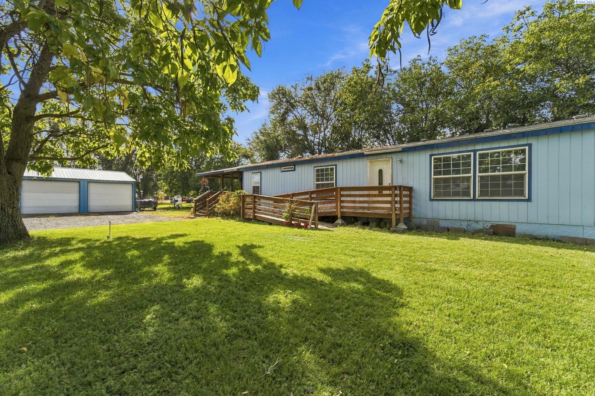 Light-blue home with a raised wooden deck and ramp, detached garage, and shady lawn.