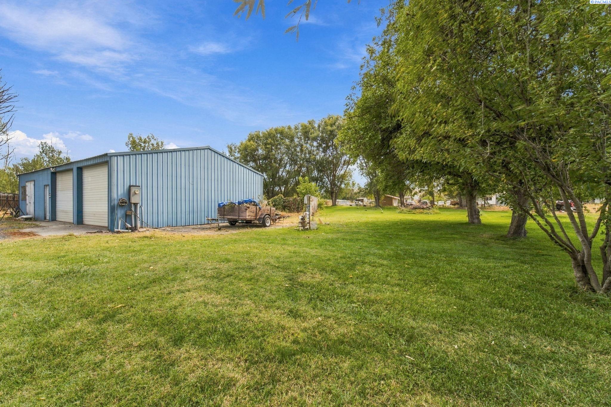 Grassy yard with mature trees and a blue metal outbuilding beside a utility trailer.