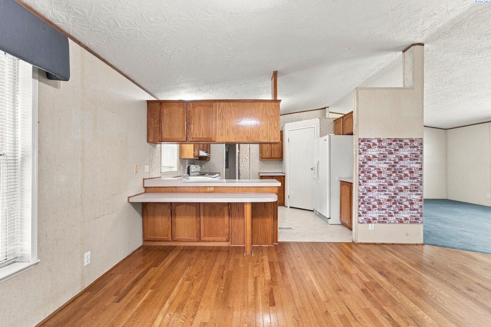 Kitchen with wood cabinets and white countertops opening onto a blue-carpeted living room.