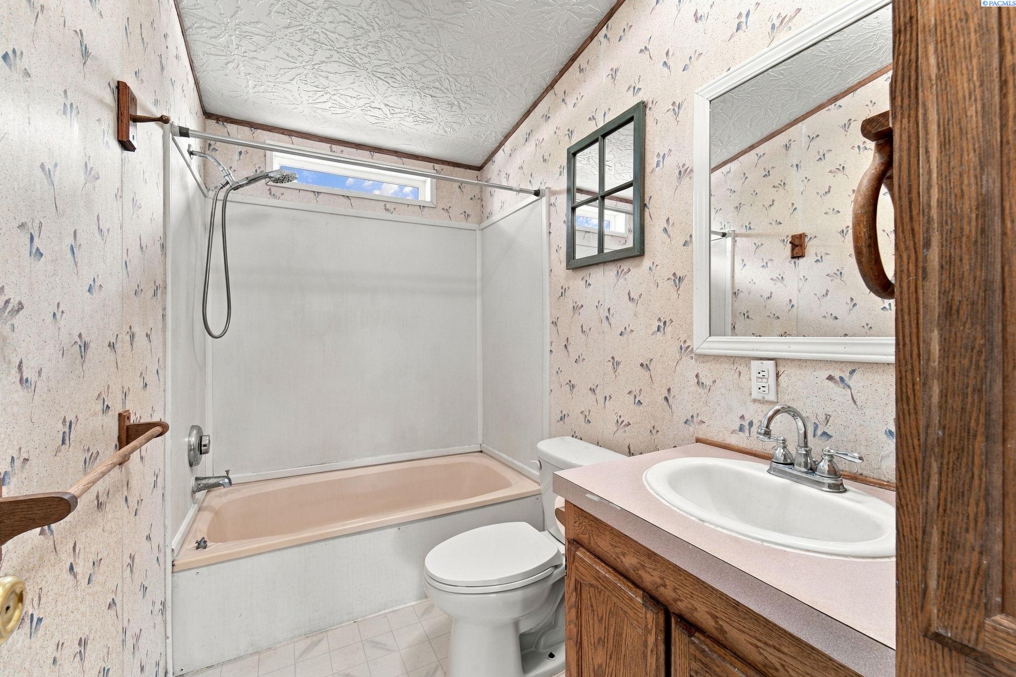 Bathroom with a tub-shower, toilet, wood vanity, and floral wallpaper.