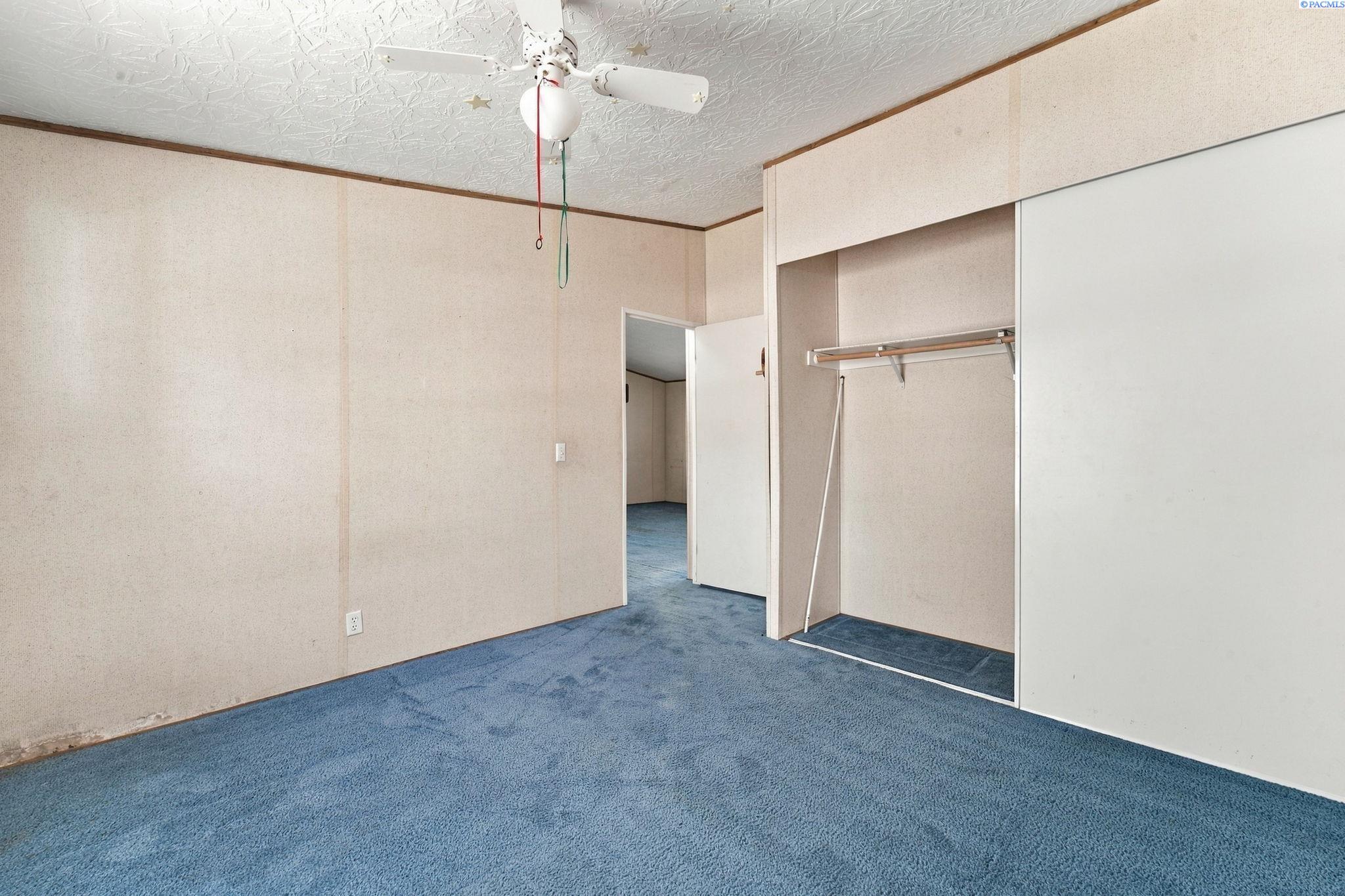 Empty bedroom with blue carpet, a ceiling fan, and an open closet with a hanging rod.