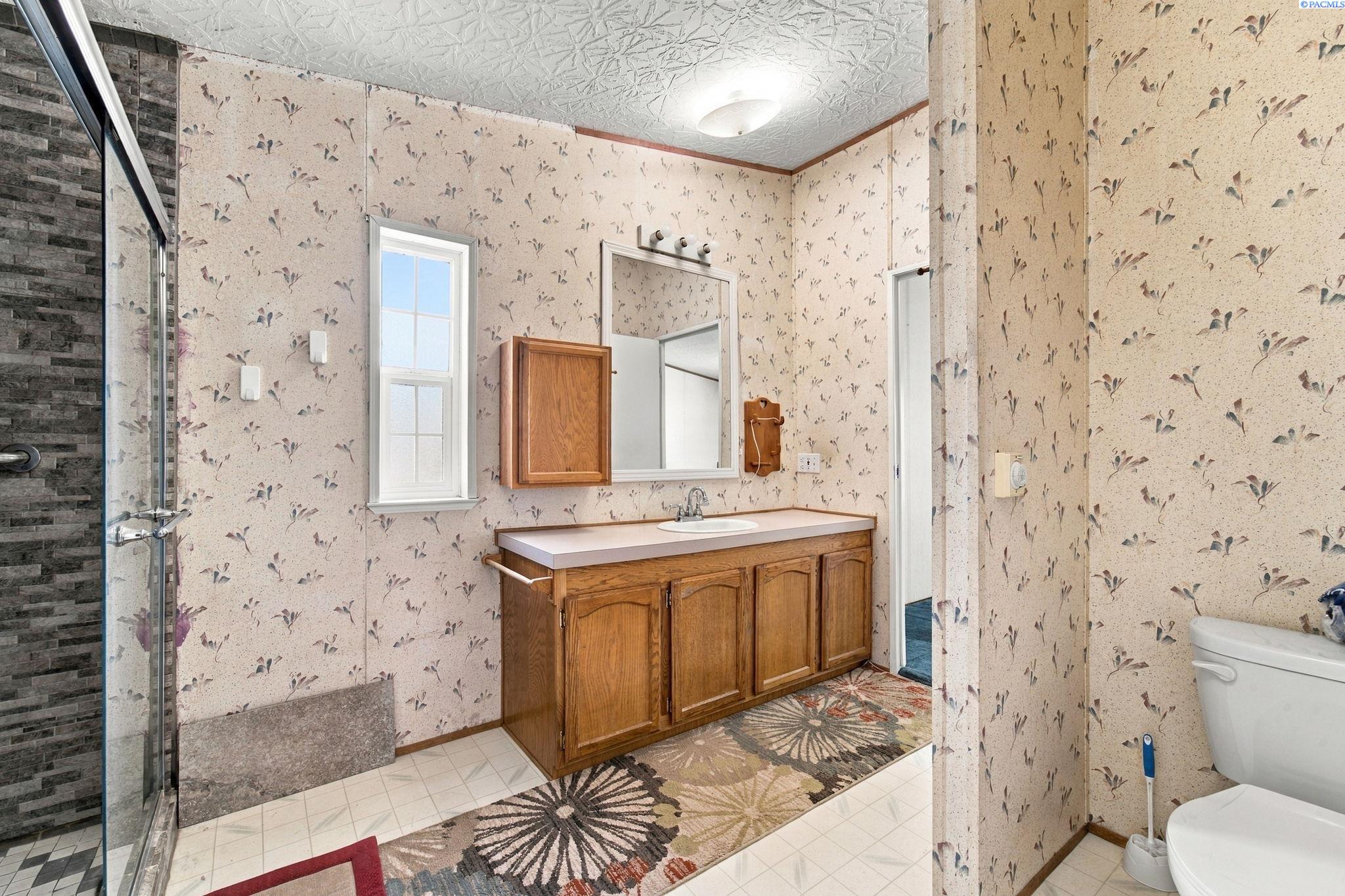Bathroom with a glass shower, floral wallpaper, and a wood vanity beneath a window.