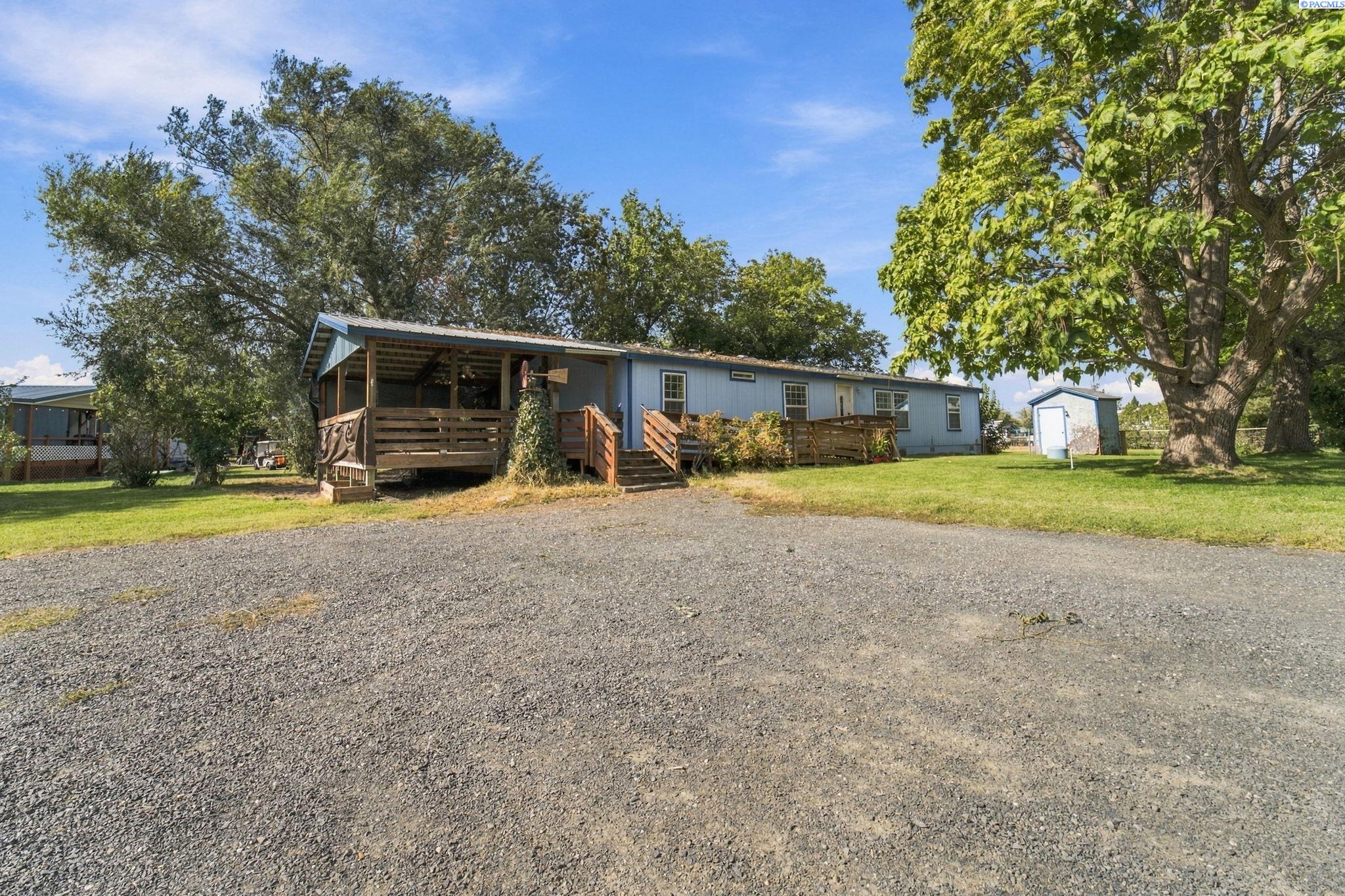 Blue single-story home with a covered wooden porch, gravel driveway, mature trees, and a small shed.