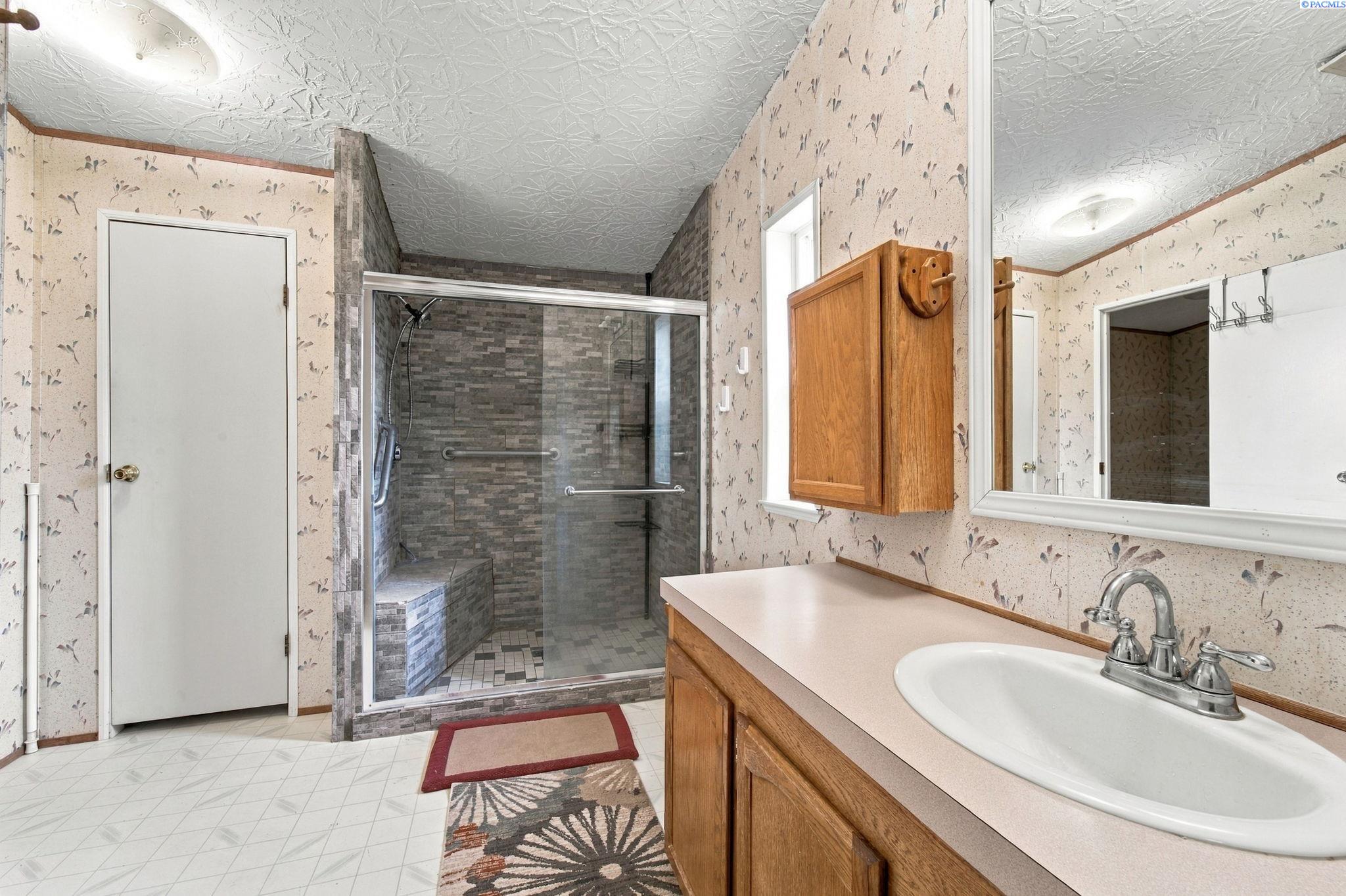 Bathroom with a gray-tiled walk-in shower, built-in seat, grab bars, and long wooden vanity.