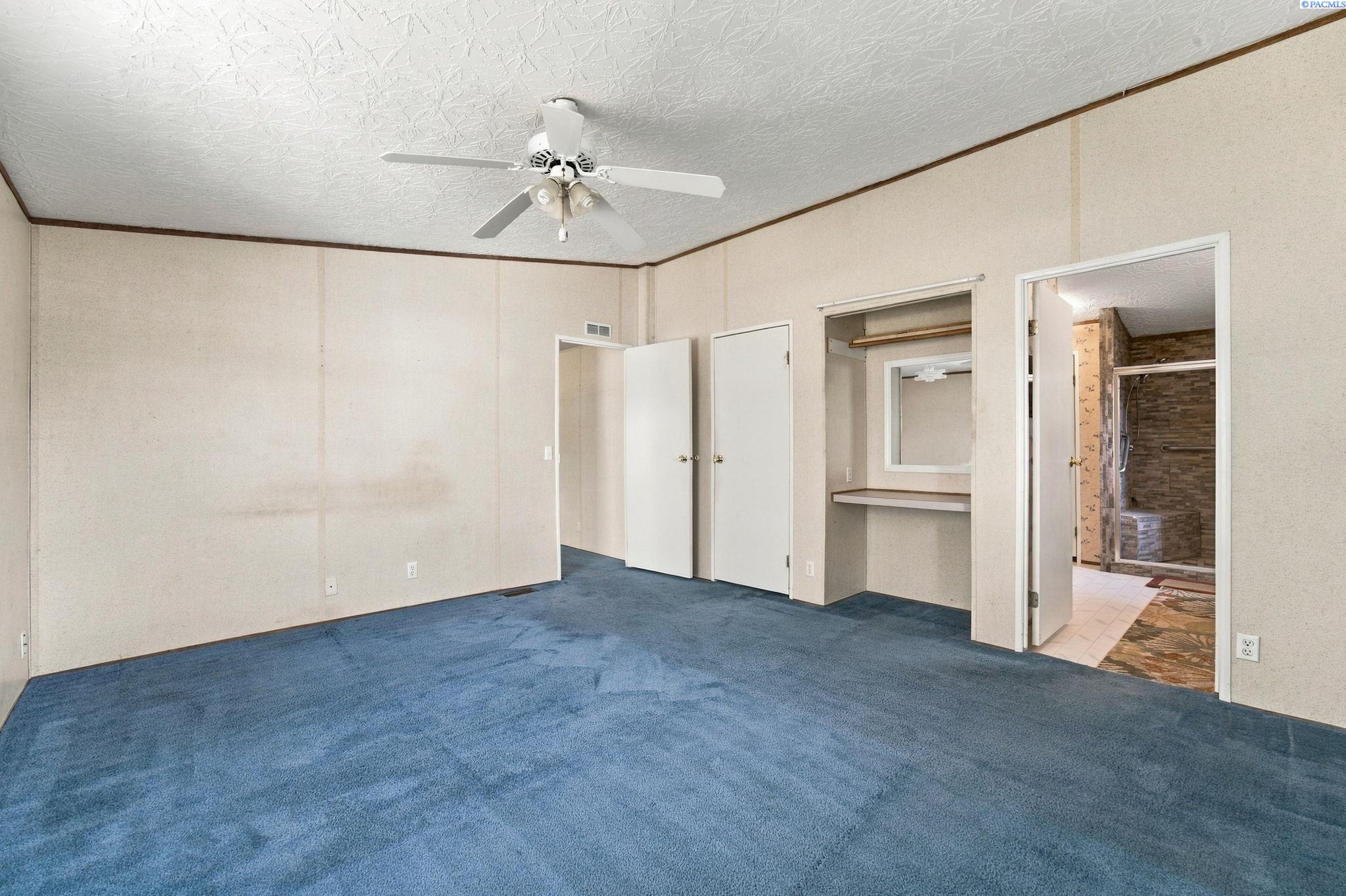 Vacant bedroom with blue carpet, ceiling fan, vanity nook, and an open doorway to a tiled bathroom.