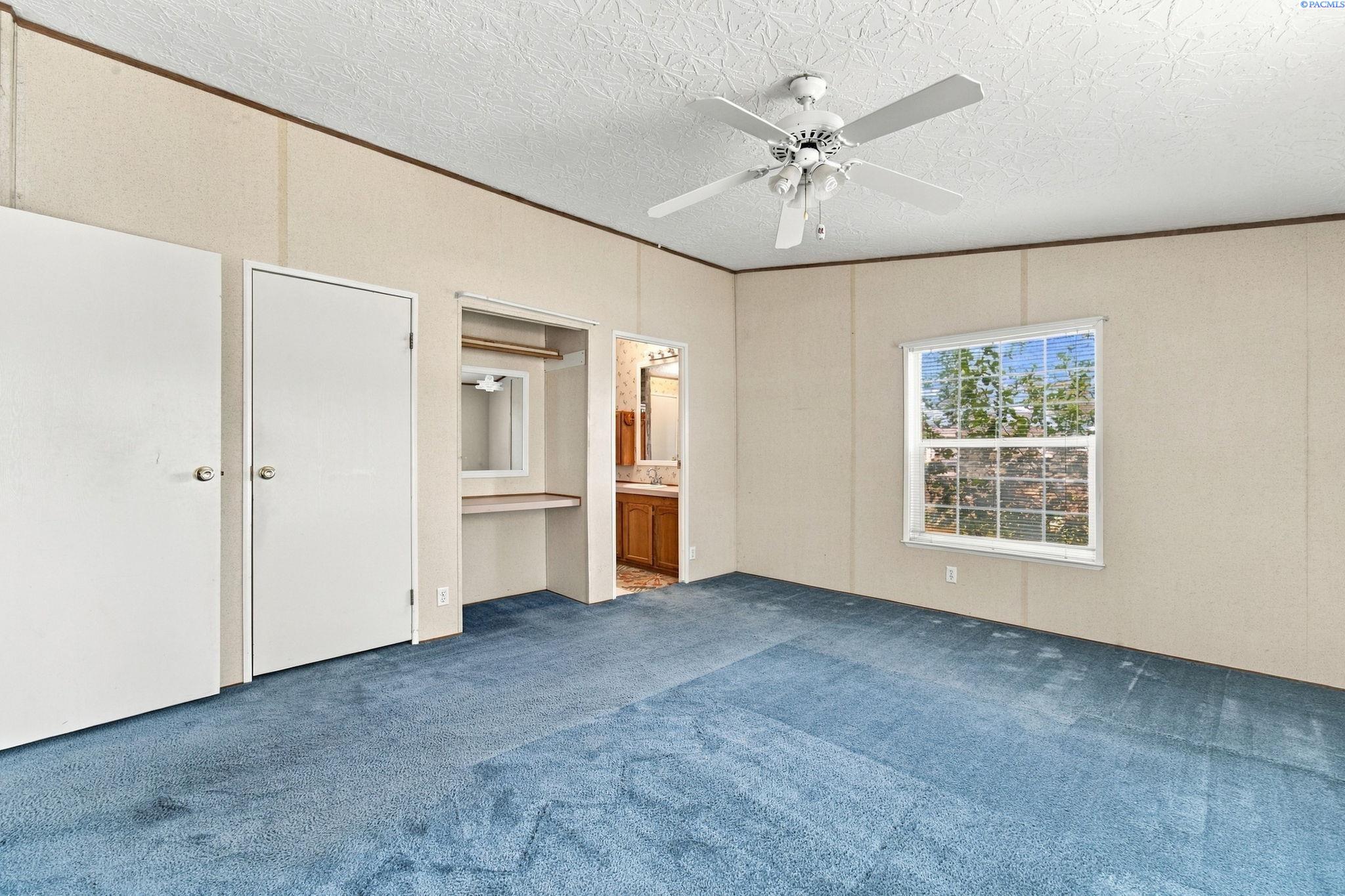 Empty bedroom with blue carpet, a ceiling fan, and an open dressing area leading toward a bathroom.
