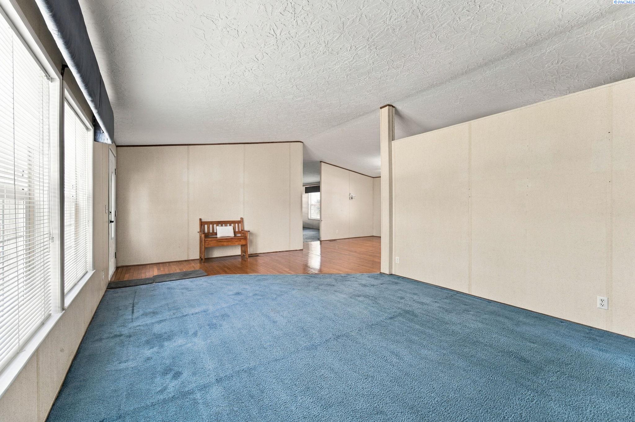 Living room with blue carpet, large windows, and a wooden bench beside adjoining hardwood flooring.