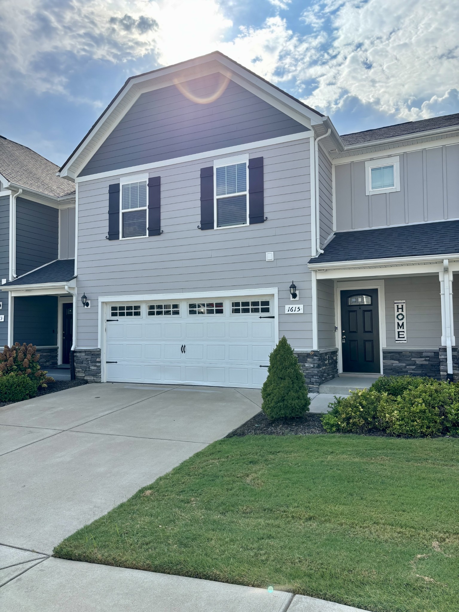 Gray two-story townhouse with dark shutters, white double garage door, and landscaped front lawn.