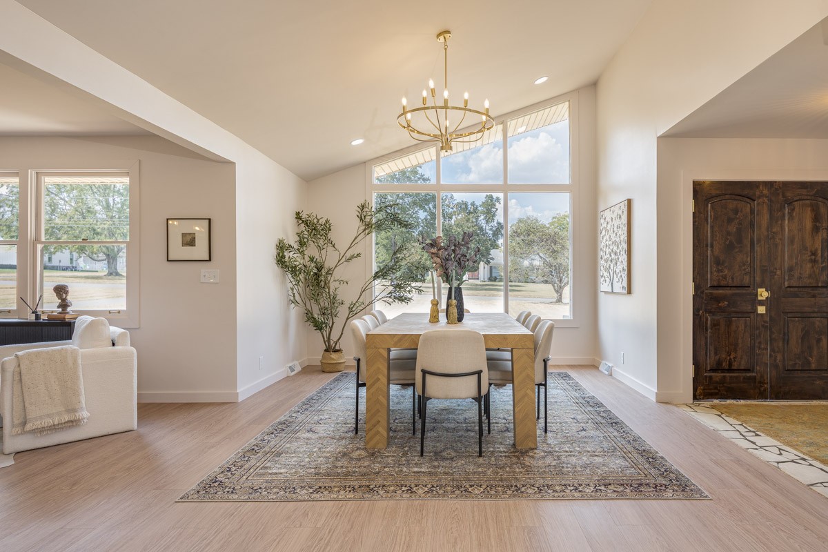 Dining area with a chandelier, large windows, and a wood table on a patterned rug.