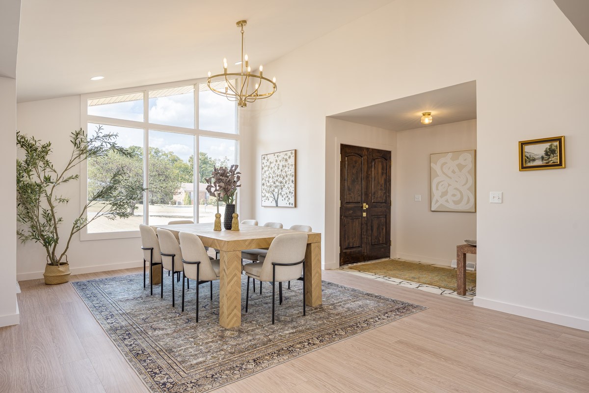 Dining room with a wood table, upholstered chairs, tall windows, and brass chandelier.